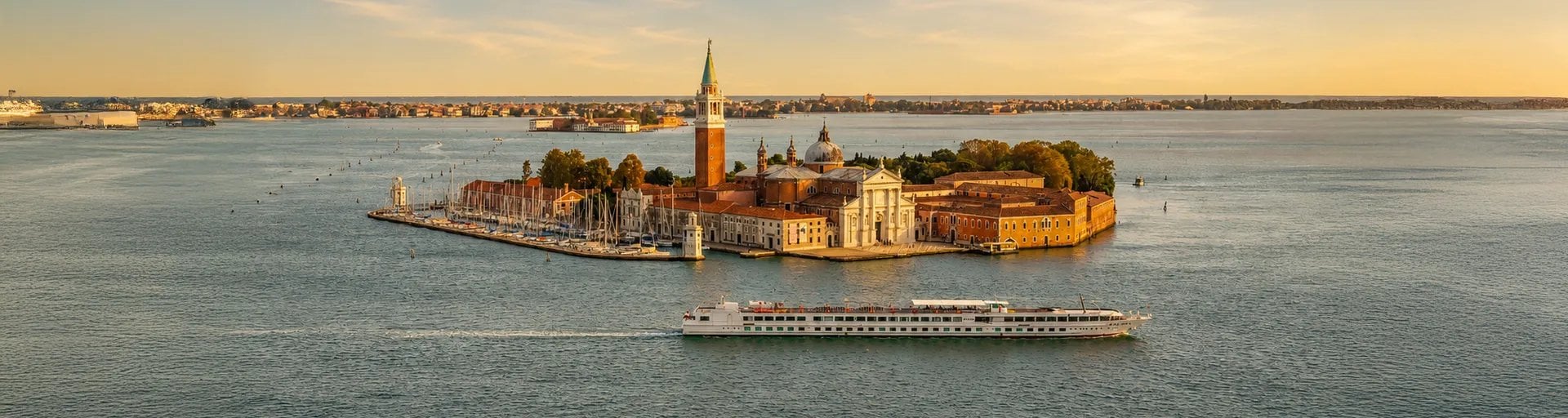 Île de San Giorgio Maggiore à Venise avec basilique, campanile et navire de croisière fluviale sur le lagon au coucher du soleil.